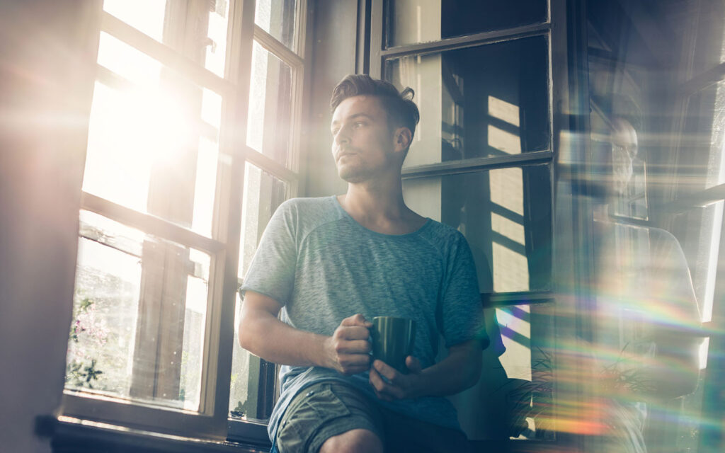Man staring out sunny window with coffee, in recovery from addiction.